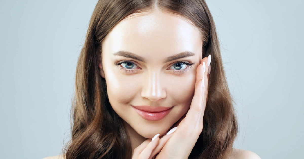 Close-up of a young woman with wavy brown hair and glowing skin, representing Metabolic Detoxification in Boynton Beach, FL, against a soft blue-gray background.